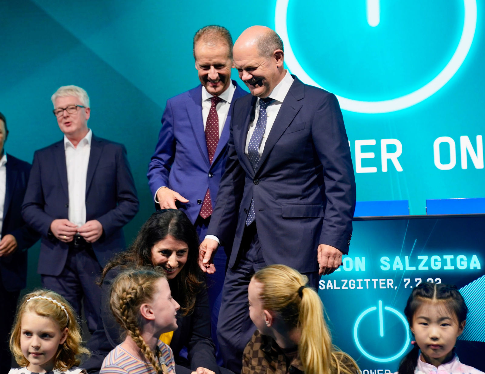 Bundeskanzler Olaf Scholz und weitere Anwesende, darunter VW-Manager, nehmen an einer Veranstaltung zur Eröffnung der Batteriefabrik „Salzgiga“ in Salzgitter teil. Scholz lächelt und beugt sich zu einer Gruppe von Kindern, die in der ersten Reihe sitzen. Im Hintergrund ist eine große LED-Wand mit dem Schriftzug „POWER ON“ und „SALZGIGA“ zu sehen.