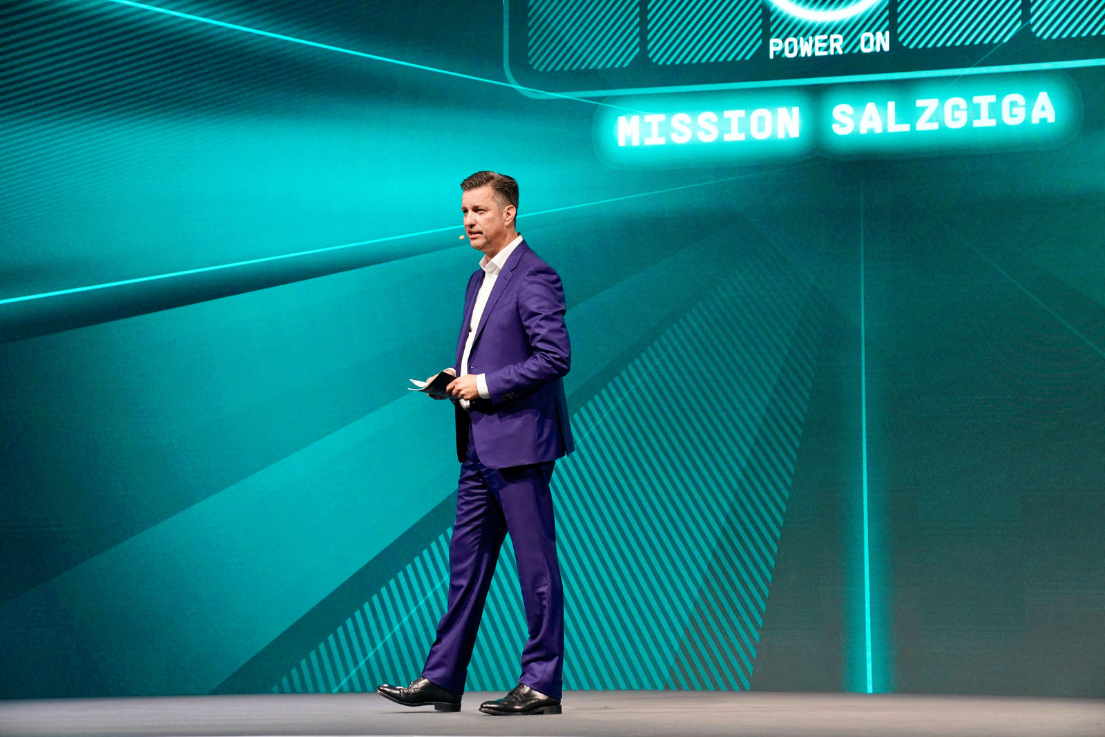 A speaker on stage at the "Mission Salzgiga" event on July 7, 2022, in Salzgitter. The speaker is wearing a blue suit and standing in front of a glowing screen projection of the event's logo.