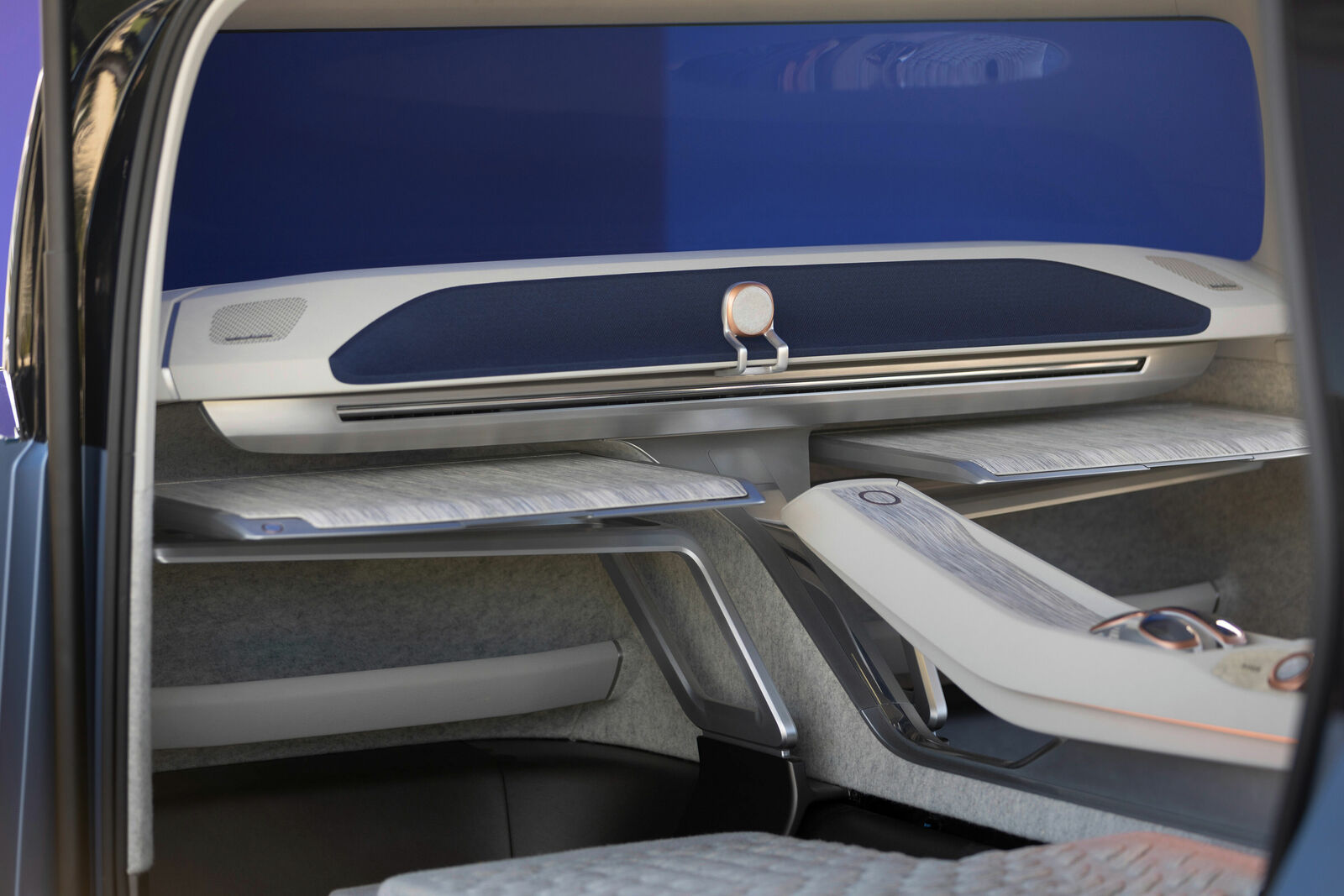 Ein futuristisches Fahrzeuginneres mit einem modernen, minimalistischen Design. Zu sehen sind stilvolle, ausklappbare Tische mit einer Holzoptik und geschwungenen Oberflächen.