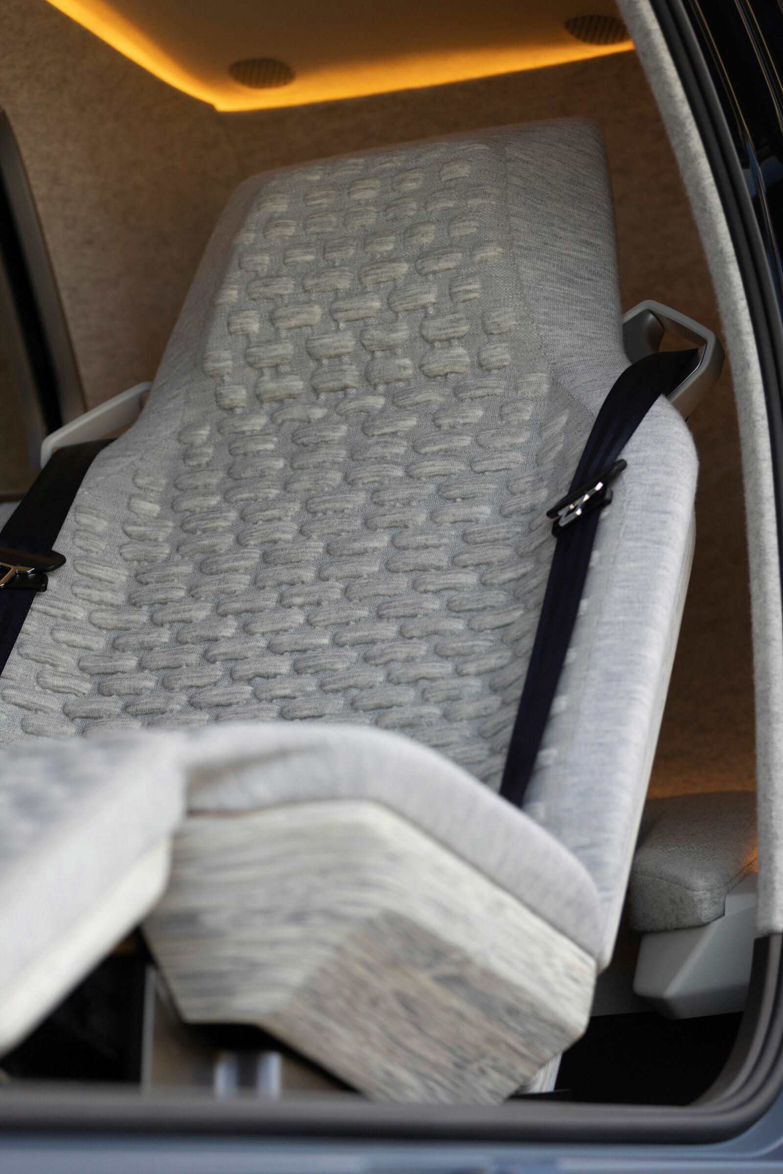 Detailaufnahme eines futuristischen Autositzes mit strukturierter Stoffpolsterung, Sicherheitsgurt und Holzoptik-Elementen. Sanfte Ambientebeleuchtung im Hintergrund.