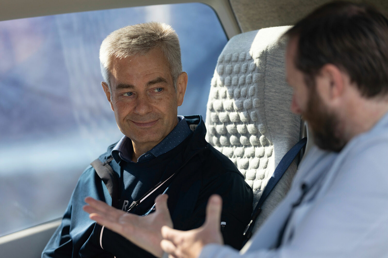 Two men are seated next to each other in a modern vehicle or simulator with cushioned seats. The man on the left is smiling and listening attentively, while the other speaks with expressive hand gestures. The scene appears familiar and focused.