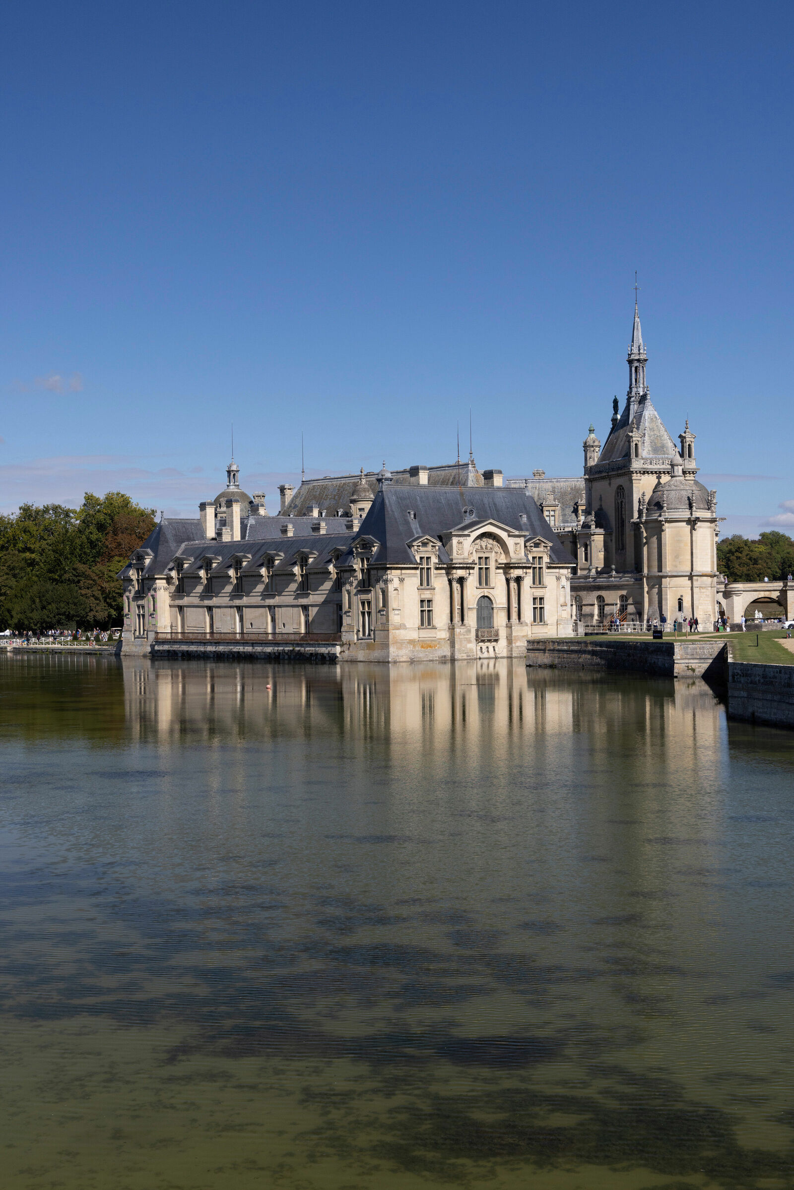 The Château de Chantilly, a historic castle in France, is reflected in the water of a surrounding lake. The sky is blue, and visitors are walking along the pathways.