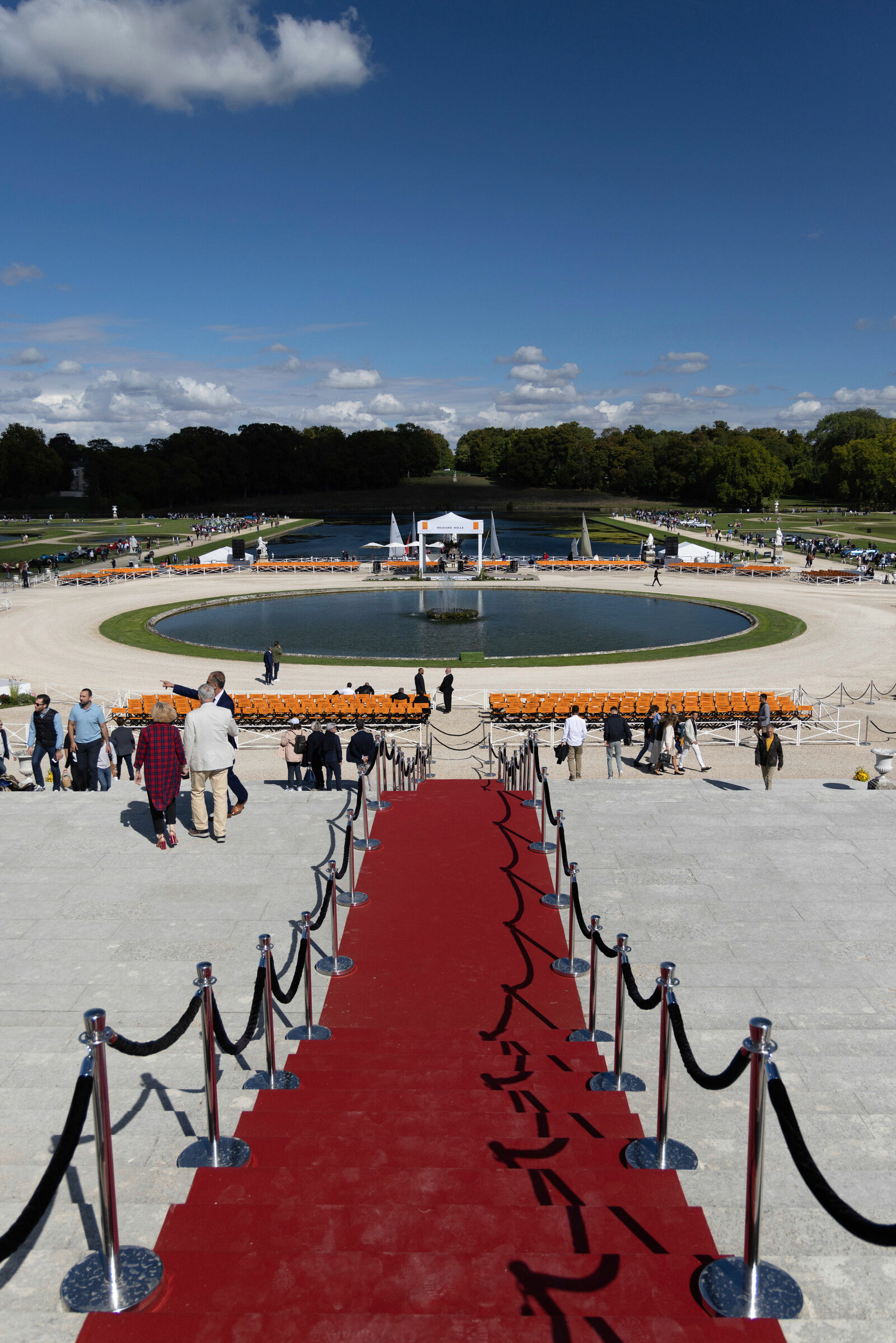 A red carpet runs down a wide stone staircase toward a circular pond with a small stage in the center. Around the pond, numerous orange chairs are arranged, and several guests are walking around the venue. The event is taking place in a large park under a blue sky with scattered clouds.