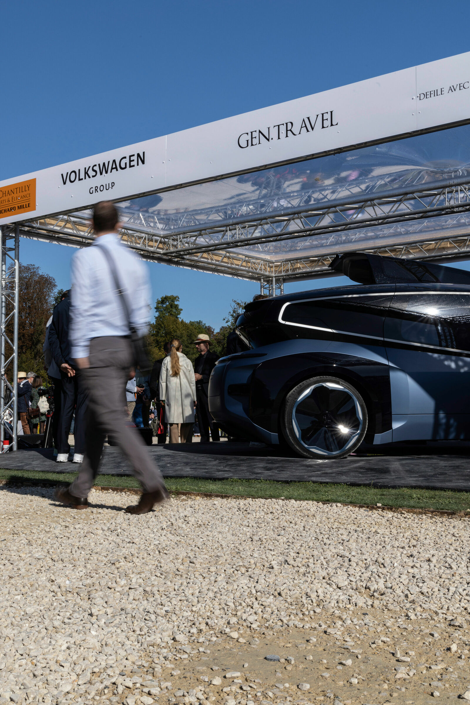 An outdoor event featuring a futuristic Volkswagen concept car under a covered exhibition area labeled "GEN.TRAVEL."