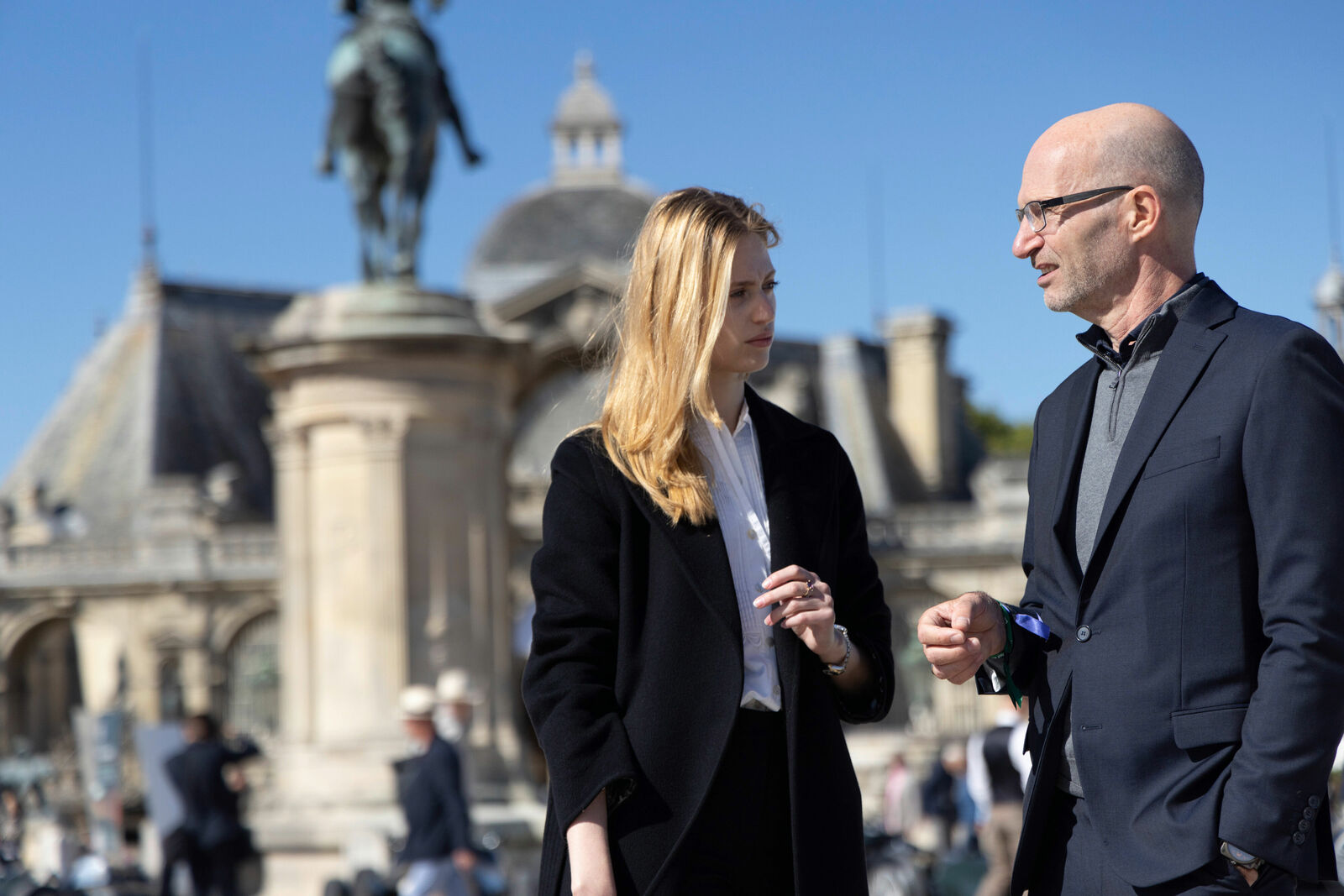 A man and a woman in elegant attire are having a conversation outdoors. The man wears a dark suit with glasses and holds a small object in his hand. The woman wears a black coat over a white shirt.