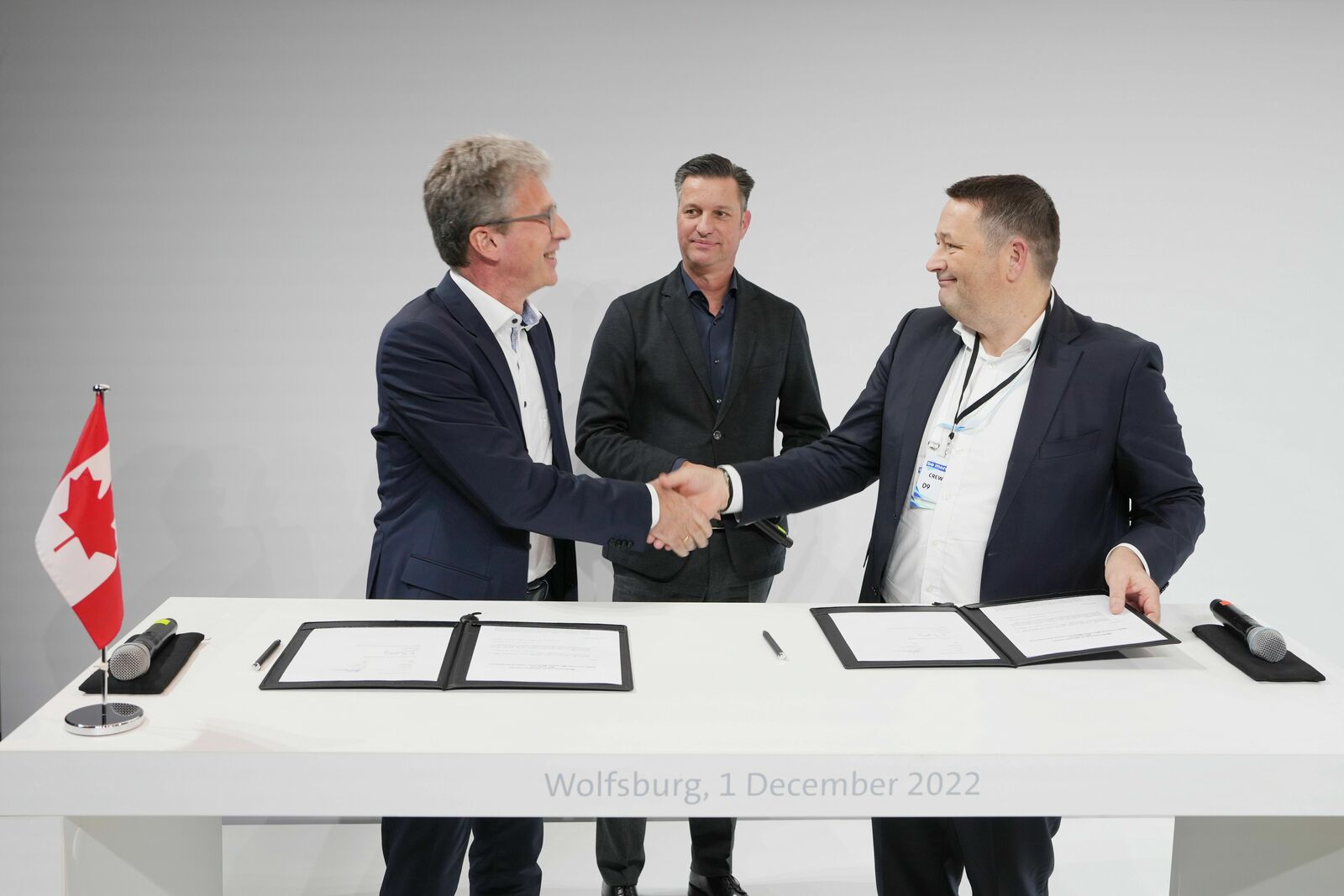 Two men in suits shake hands at a white table with signed documents, while a third man observes in the background. A small Canadian flag is placed on the table, and the inscription “Wolfsburg, 1 December 2022” is visible.