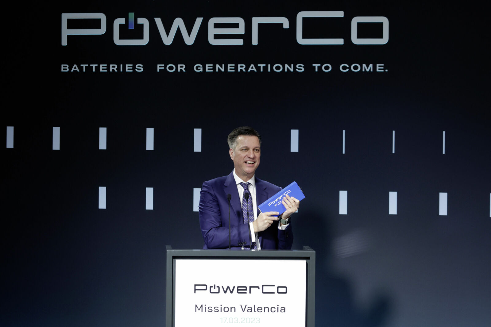 A man in a dark suit and purple tie stands behind a podium labeled "PowerCo Mission Valencia 17.03.2023." He holds a small blue box with the PowerCo logo and smiles.