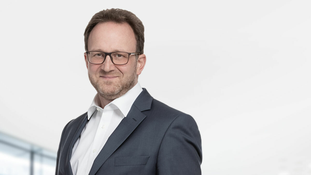 The picture shows Wolfgang Fueter against a light background. He is wearing a suit, shirt and glasses.