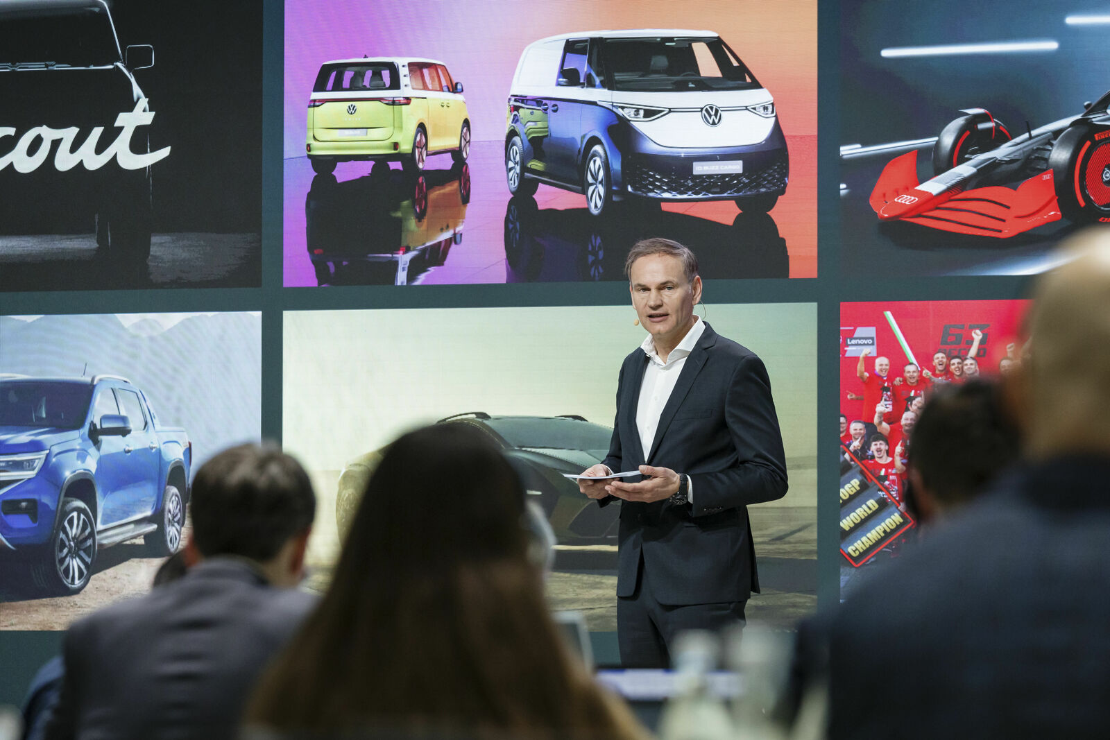 A man in a dark suit stands on a stage, giving a presentation to an audience. Behind him, a large screen displays images of various vehicles, including the Volkswagen ID.Buzz, an SUV, a race car, and a concept vehicle. The audience in the foreground listens attentively.