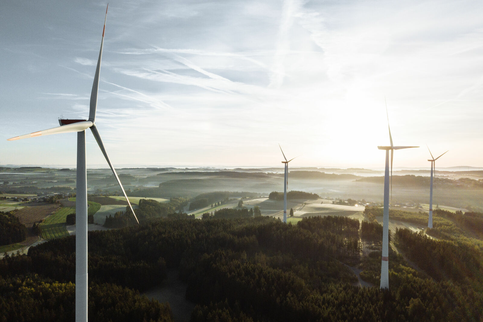 Aerial view of four wind turbines standing in a forested landscape. The sun rises over the rolling hills, creating an atmospheric lighting effect.