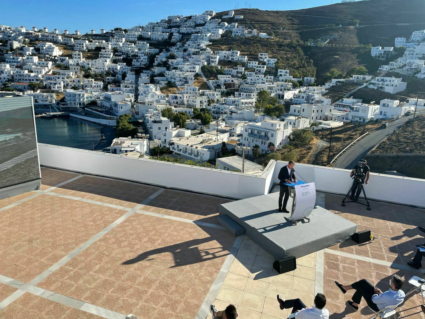 Ein Redner steht auf einer kleinen Bühne mit Rednerpult auf einer Terrasse mit Blick auf eine griechische Inselstadt mit weißen Häusern an einem Hügel. Das Publikum sitzt entspannt auf Stühlen, während Kameras die Szene aufnehmen.