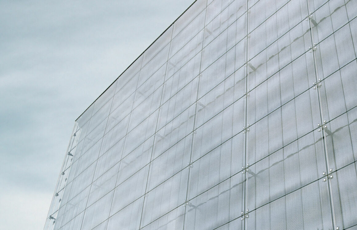 Minimalist architectural shot of a modern building with a glass or metal facade. The surface reflects the sky, creating a geometric pattern of lines and fastening points. The perspective is slightly tilted upward, emphasizing the building’s height.