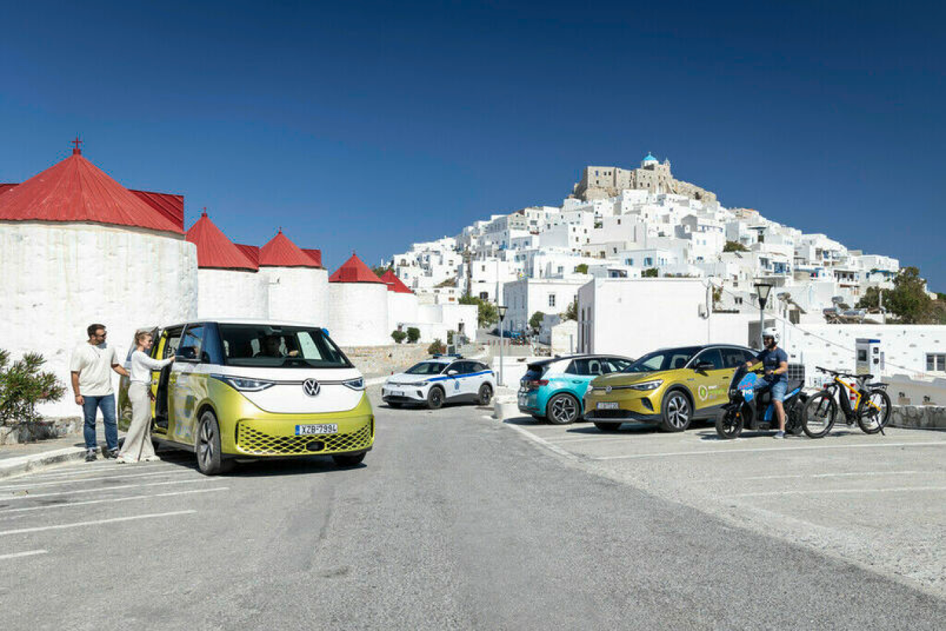 Eine bunte Gruppe von Fahrzeugen und Menschen auf einer Straße auf einer griechischen Insel. Im Vordergrund ein gelber Volkswagen Bus