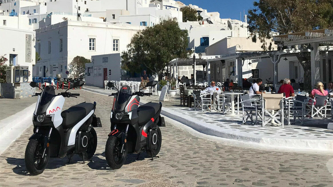 Zwei Elektromotorräder auf einer gepflasterten Straße vor einem Café in einer weißen, griechischen Stadt.