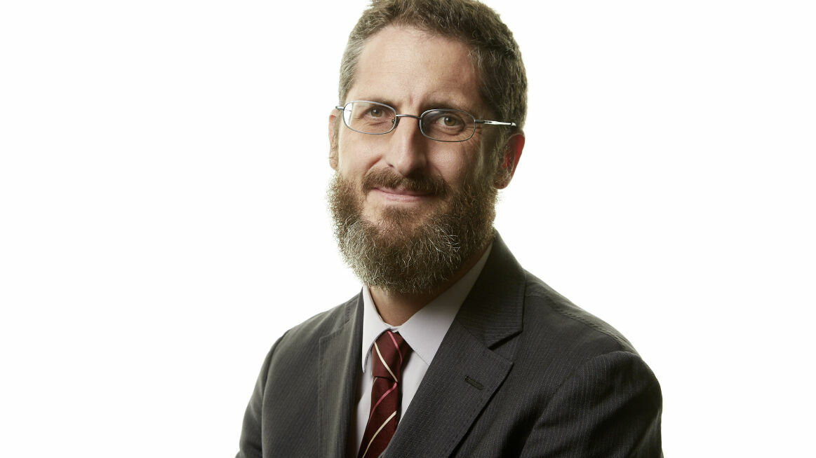 A man with glasses and a beard is wearing a suit and tie, smiling slightly at the camera.