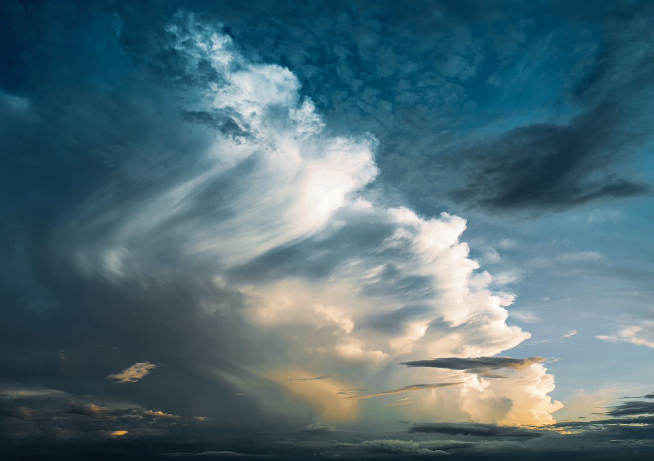 Eine beeindruckende Wolkenformation am Himmel, die sich in verschiedenen Blautönen und warmen goldenen Lichtspiegelungen erstreckt. Die Wolken wirken dramatisch und dynamisch, als würden sie sich in Bewegung befinden.