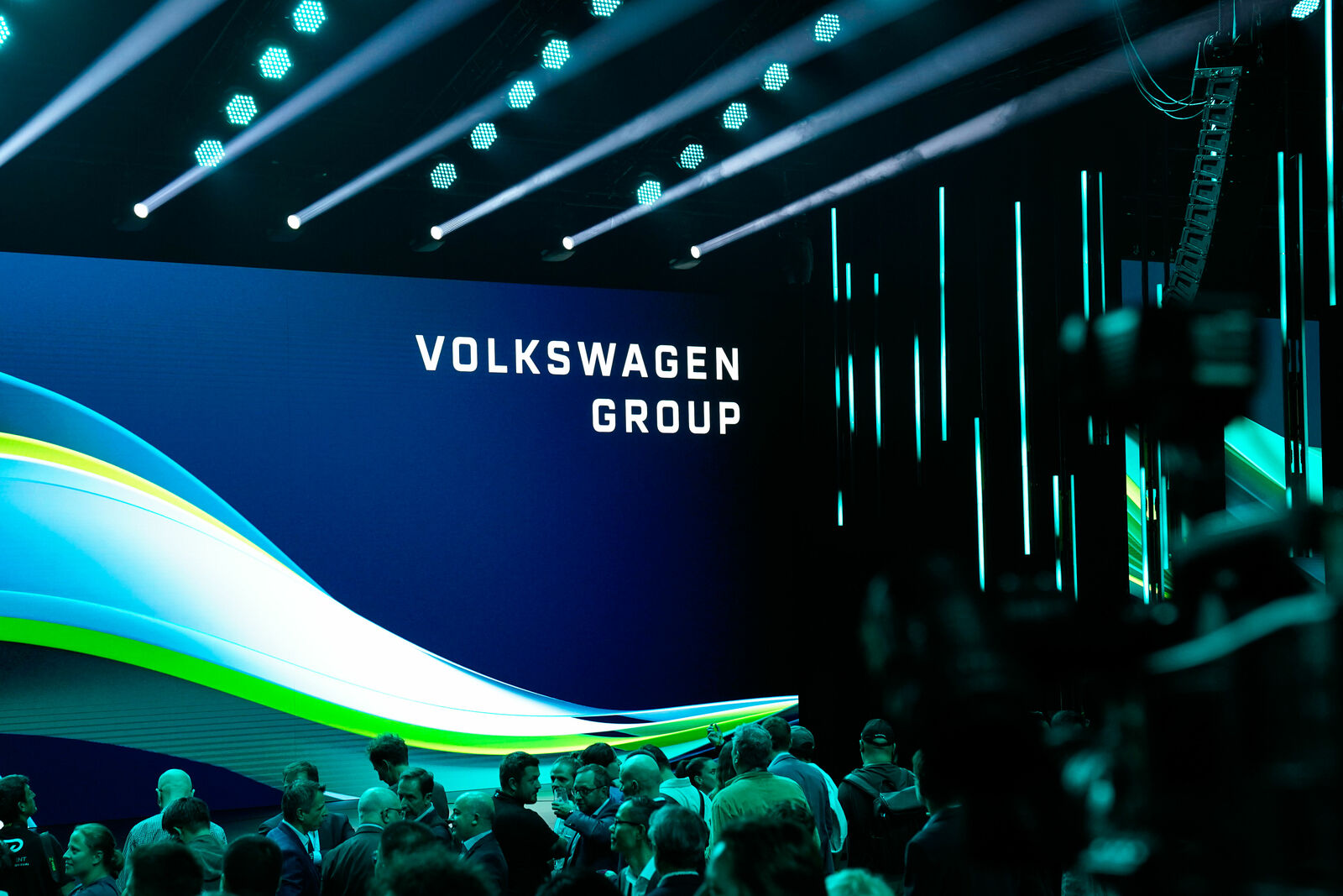 Eine Veranstaltung mit dem Volkswagen Group Logo auf einer großen LED-Leinwand. Die Bühne ist in Blau- und Grüntönen beleuchtet, mit dynamischen Lichtstreifen im Hintergrund. Im Vordergrund stehen zahlreiche Teilnehmer, die sich unterhalten. Kameras und Technik sind ebenfalls sichtbar, was auf eine professionelle Veranstaltung oder Präsentation hinweist.