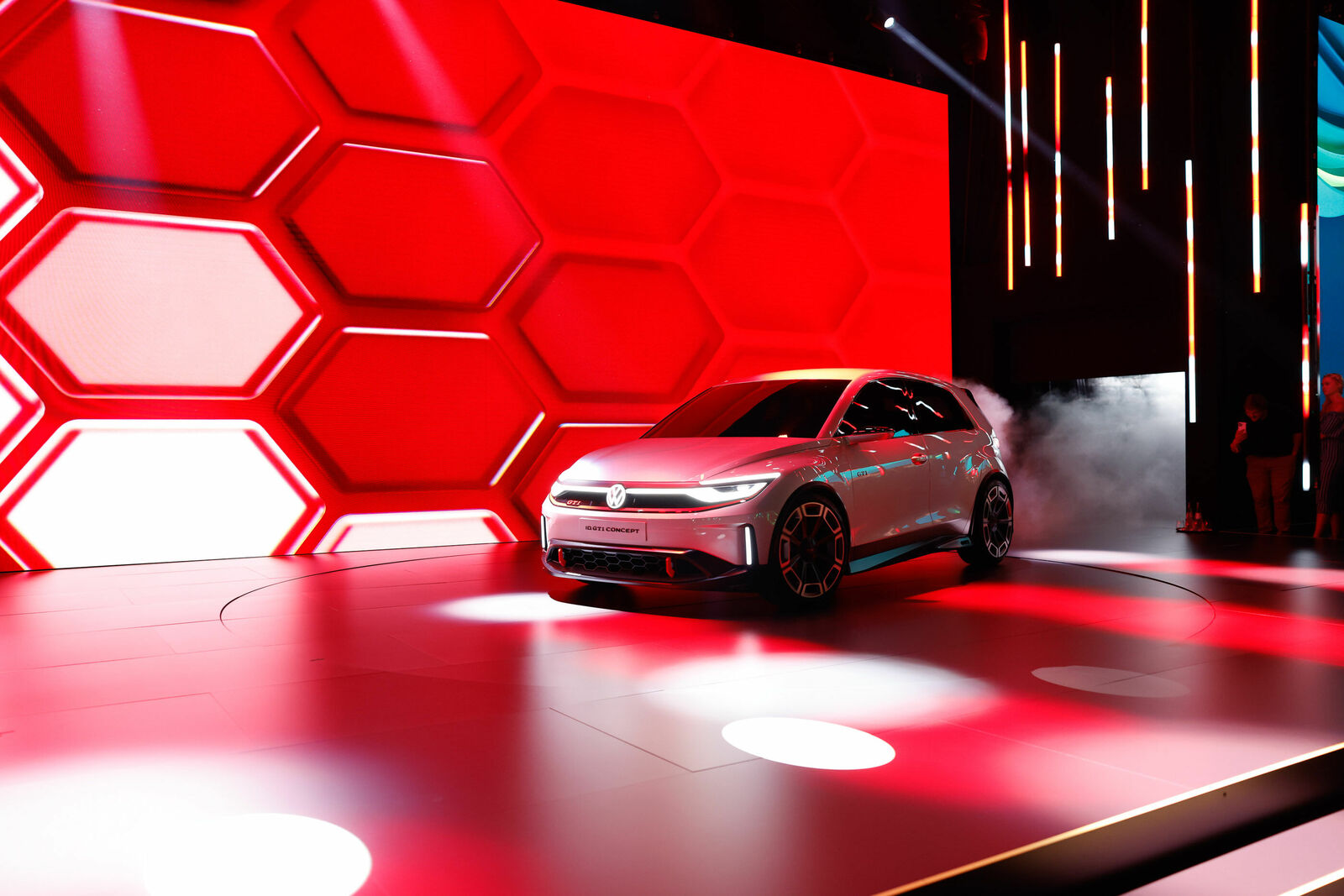 A futuristic silver Volkswagen concept car is showcased on a stage with a red honeycomb-patterned background. Spotlights and lighting effects create a dynamic atmosphere, while fog rises from the vehicle area.