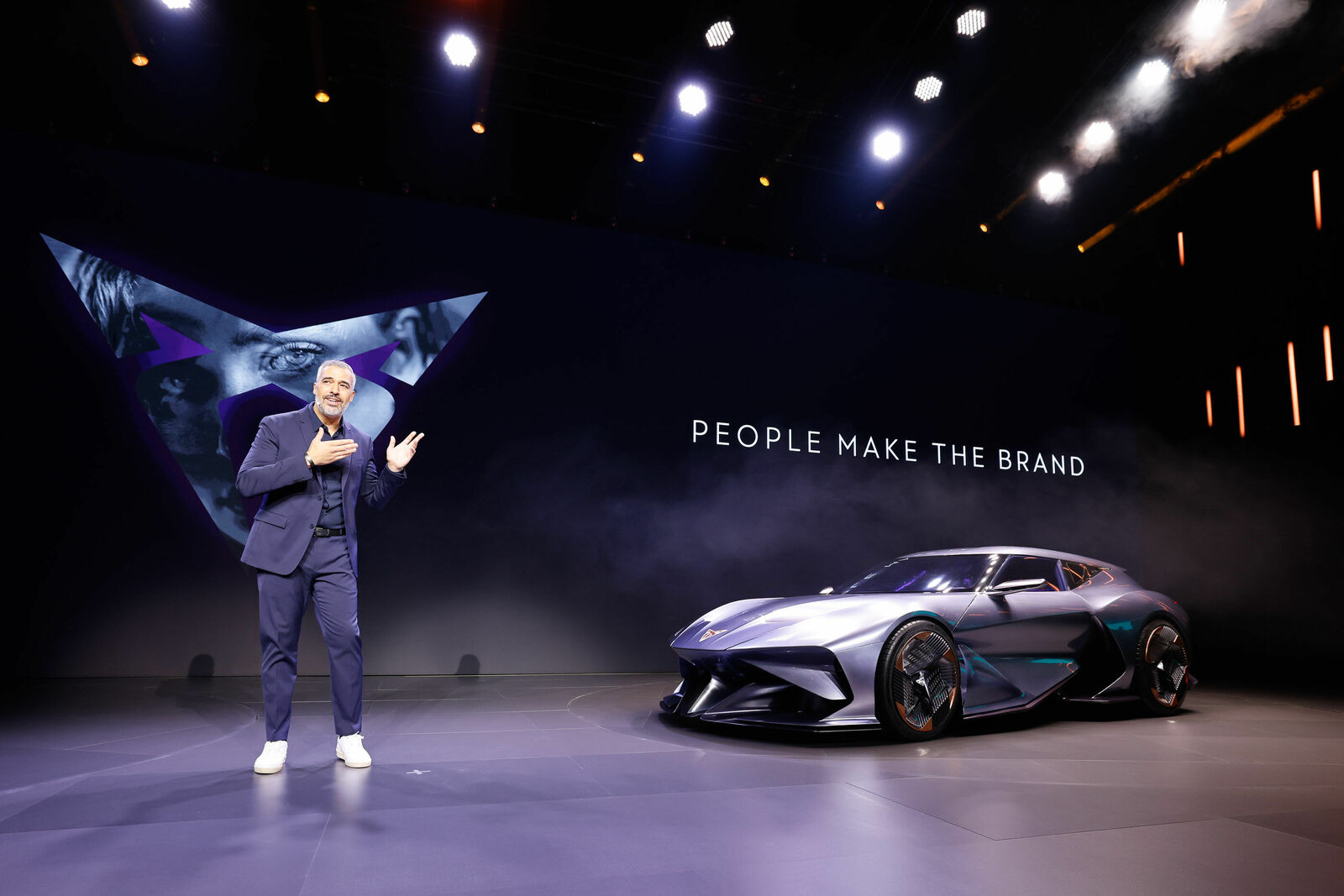A man in a dark blue suit stands on a stage, giving a presentation. Next to him is a futuristic, sporty car with sharp lines and an aerodynamic design. In the background, a large screen displays the slogan "PEOPLE MAKE THE BRAND" along with a graphic logo featuring faces and purple accents. The stage has a dark theme, illuminated by modern lighting effects.