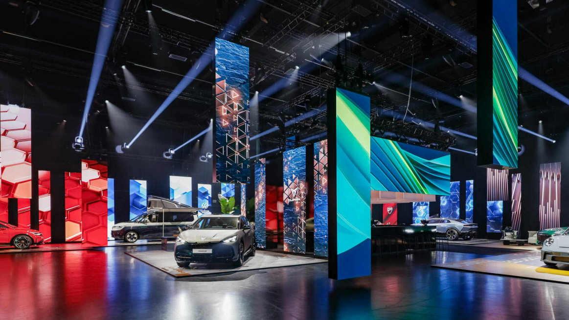 Eine Autoausstellung mit modernem Design, die mehrere Fahrzeuge auf beleuchteten Podesten präsentiert, umgeben von dynamischen, farbigen Strukturen und Säulen, die mit Grafiken verkleidet sind.