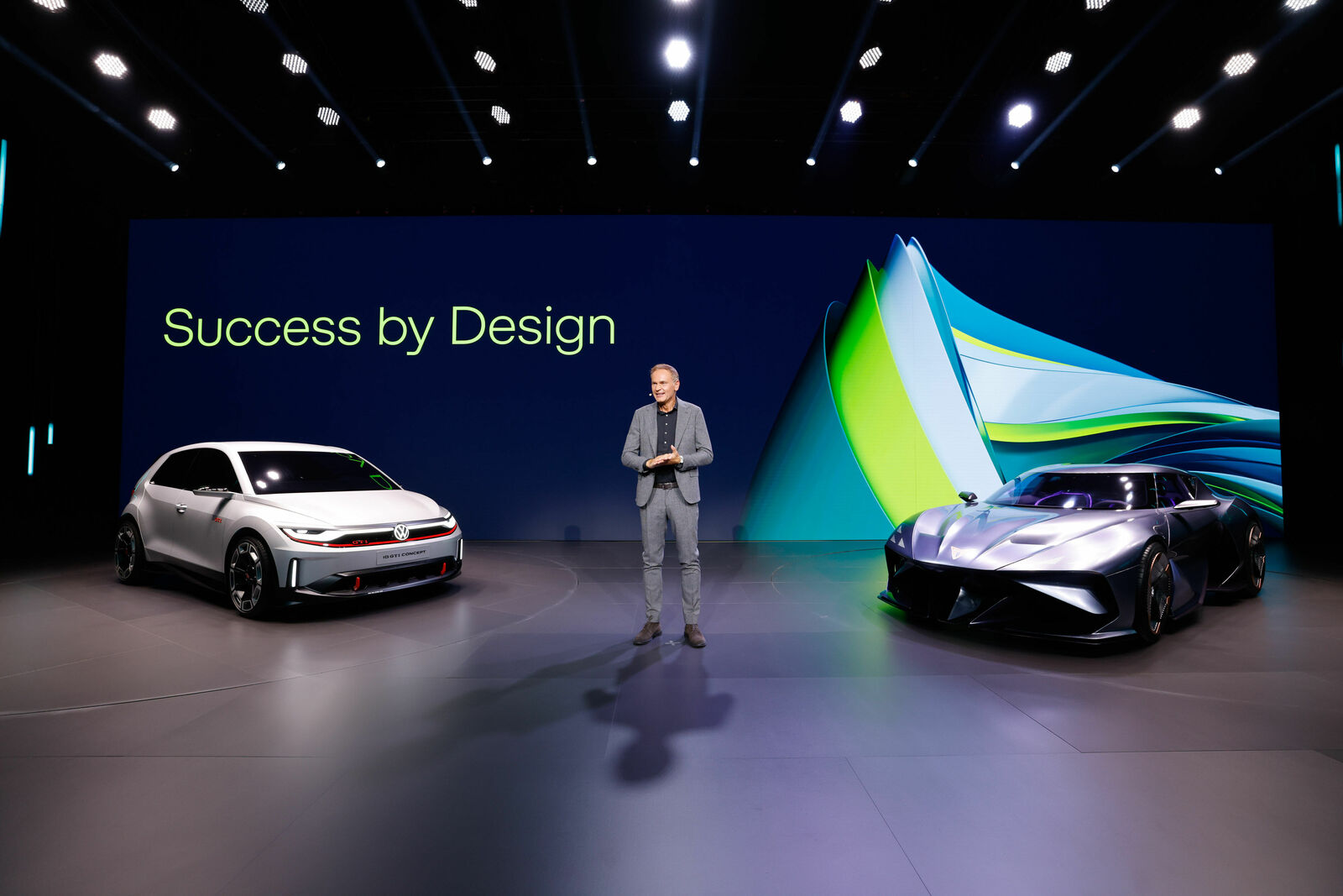 Ein Mann in einem grauen Anzug steht auf einer Bühne zwischen zwei futuristischen Volkswagen-Konzeptfahrzeugen. Hinter ihm ist eine große LED-Wand mit der Aufschrift „Success by Design“ und einer abstrakten Grafik in Blau und Grün zu sehen. Die Szene ist professionell ausgeleuchtet.