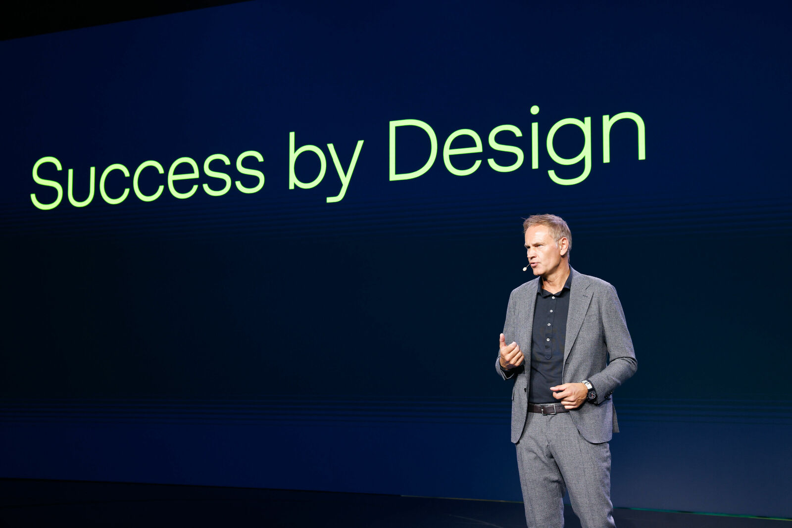 Ein Mann in einem grauen Anzug hält eine Präsentation auf einer Bühne. Hinter ihm auf einer großen Leinwand steht in leuchtend grüner Schrift „Success by Design“. Er spricht mit einem Headset-Mikrofon und gestikuliert mit einer Hand.