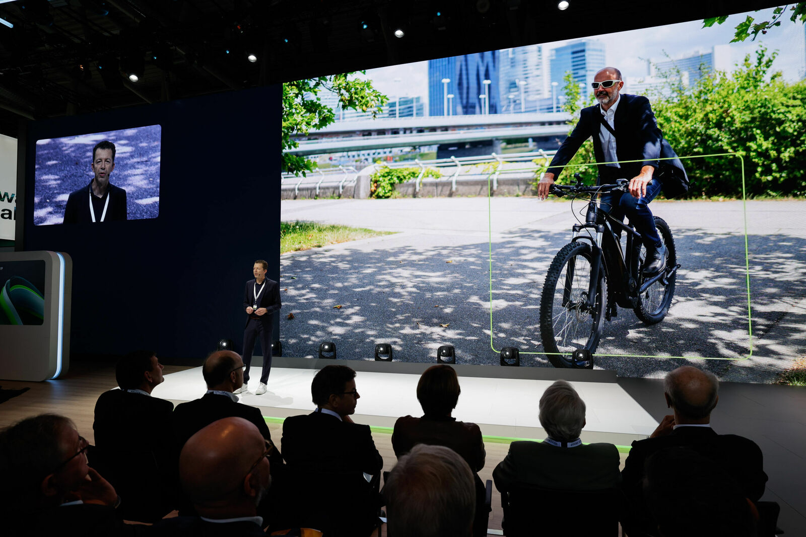 Ein Redner steht auf einer Bühne vor einem großen LED-Bildschirm, auf dem ein Mann in Anzug und Sonnenbrille auf einem E-Bike in einer urbanen Umgebung zu sehen ist. Das Publikum, bestehend aus mehreren Geschäftsleuten, verfolgt die Präsentation aufmerksam.