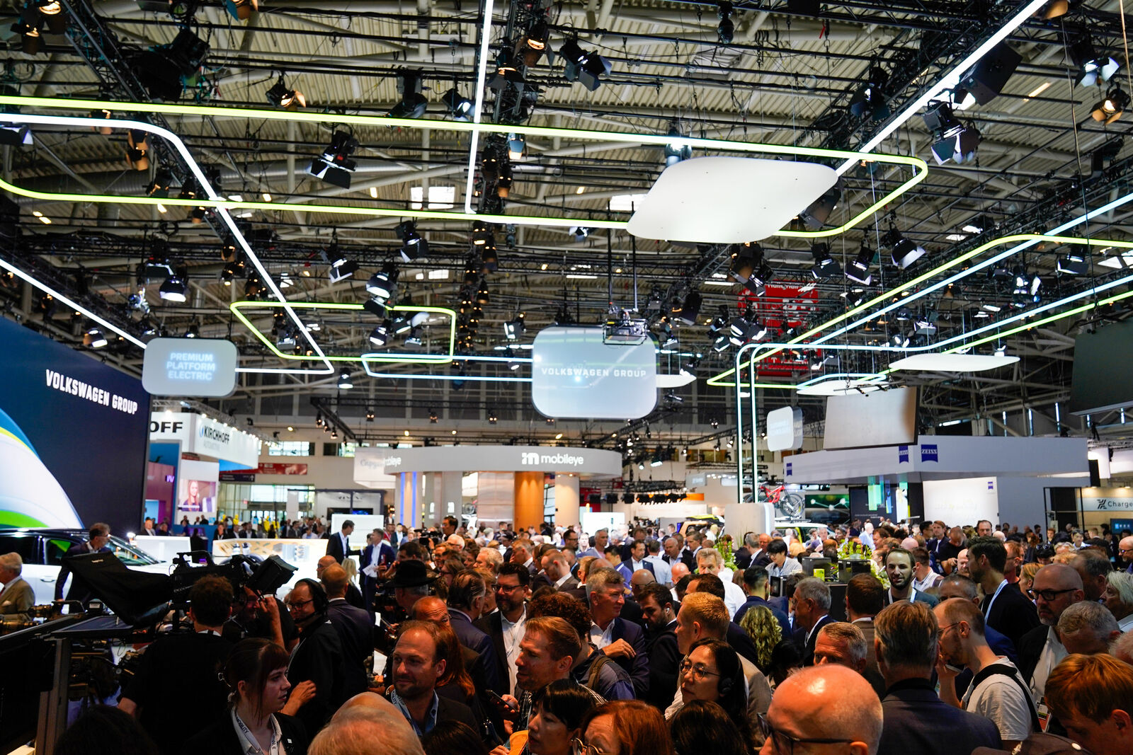 Eine große Menschenmenge besucht eine Automobilmesse mit zahlreichen Ständen und Displays. Im Vordergrund ist der Stand der Volkswagen Group zu sehen, umgeben von modernen Lichtinstallationen und Bildschirmen. Die Decke ist mit zahlreichen Leuchten und LED-Streifen ausgestattet.