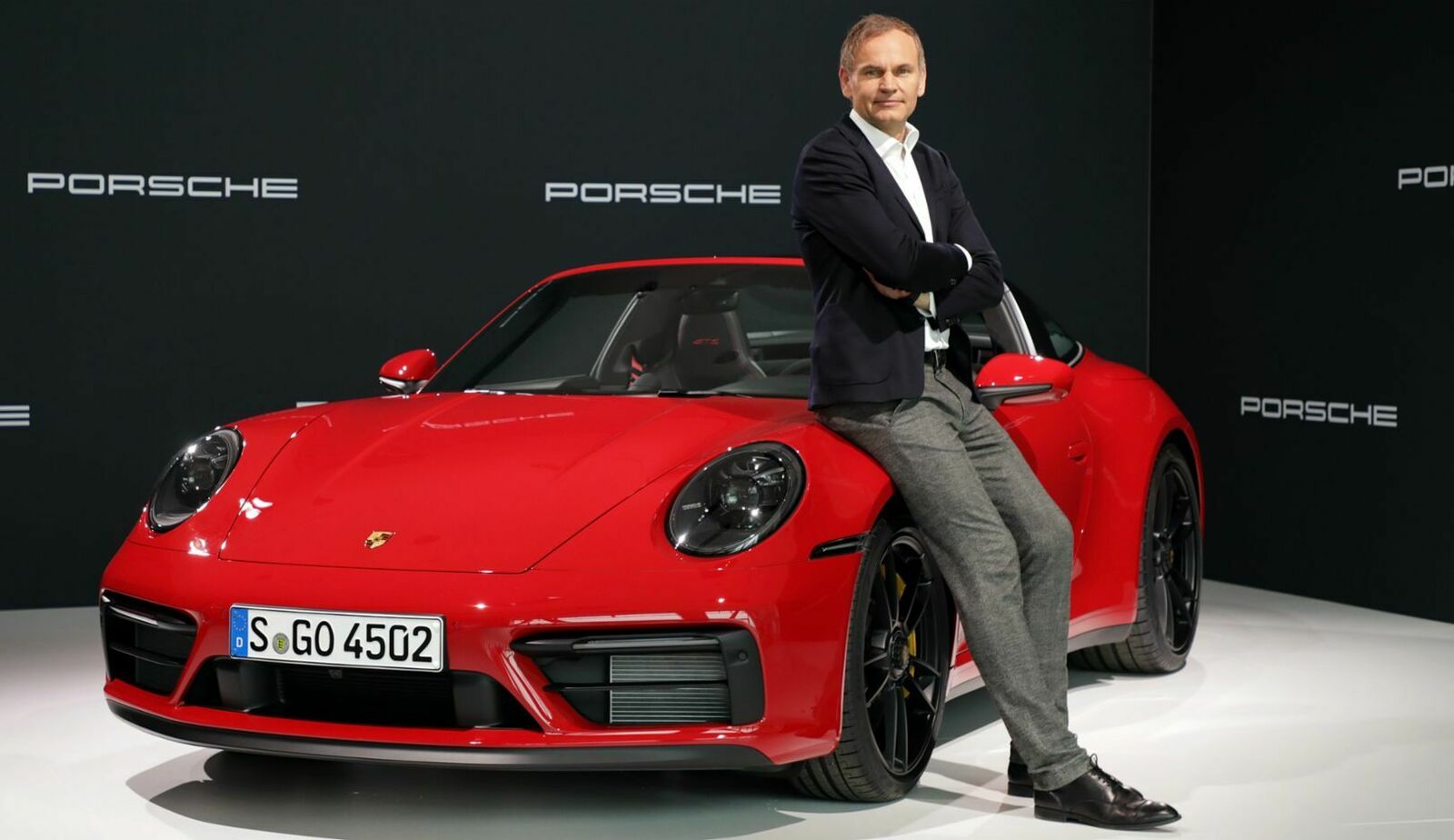 Oliver Blume leans against a red Porsche 911 and looks into the camera