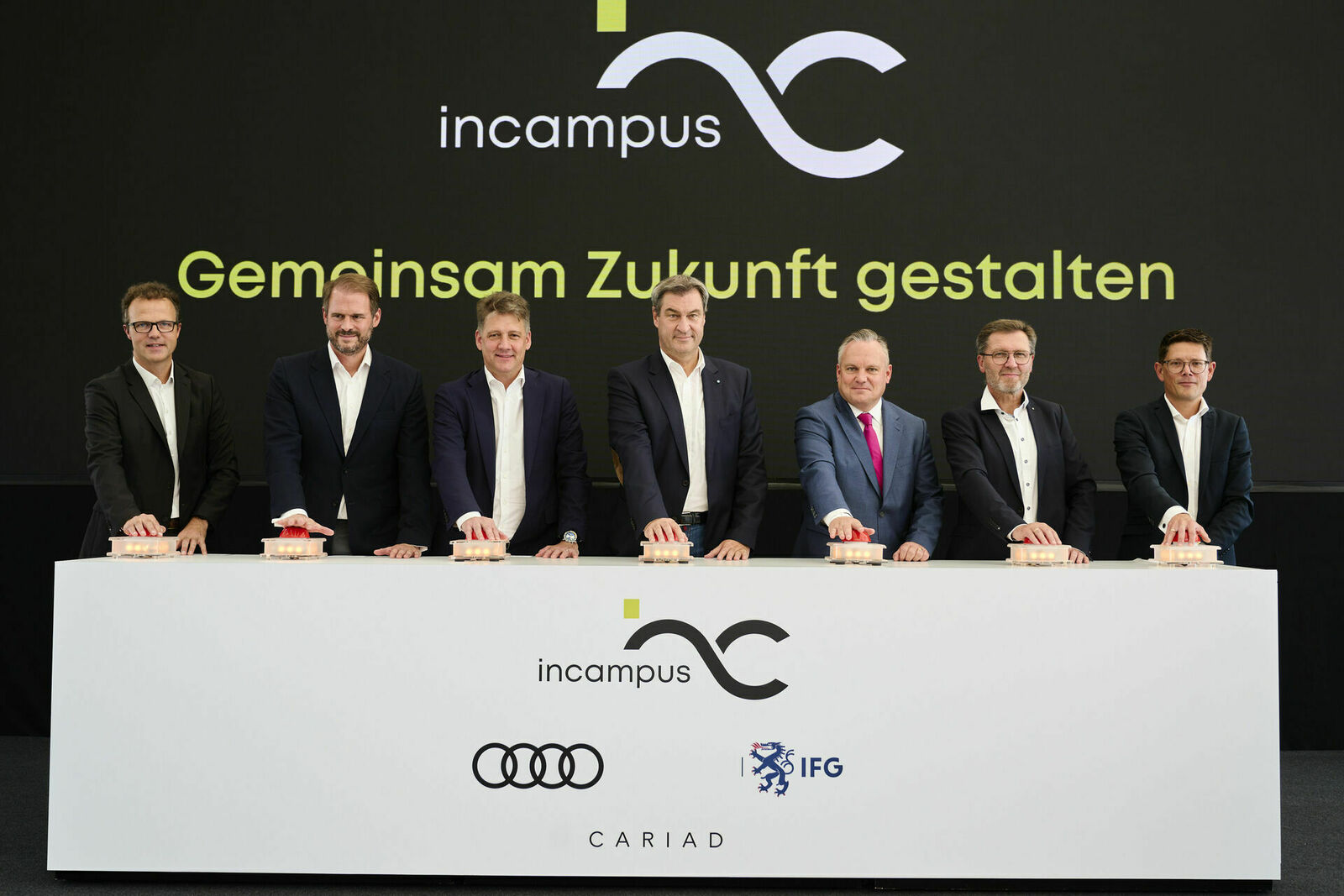 A picture of Christof Messner, Managing Director of IN-Campus GmbH and Audi Events and Services GmbH; Peter Bosch, CEO of CARIAD; Gernot Döllner, Chairman of the Board of Management of AUDI AG; Markus Söder, Bavarian Minister President; Christian Scharpf, Lord Mayor of the City of Ingolstadt; Norbert Forster, Managing Director of IN-Campus GmbH and Board Member of IFG Ingolstadt AöR and Dennis Hartmann, Overall Project Manager incampus and Project Manager New Vehicle Safety Center, standing at a lectern and placing their hands on a red button.