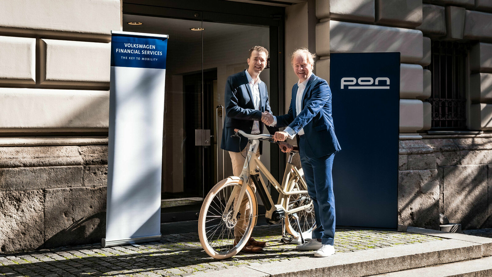 Dr. Christian Dahlheim (links), CEO der Volkswagen Financial Services AG, und Janus Smalbraak (rechts), CEO von Pon Holdings stehen zusammen vor eunem Fahrrad und geben sich die Hand.