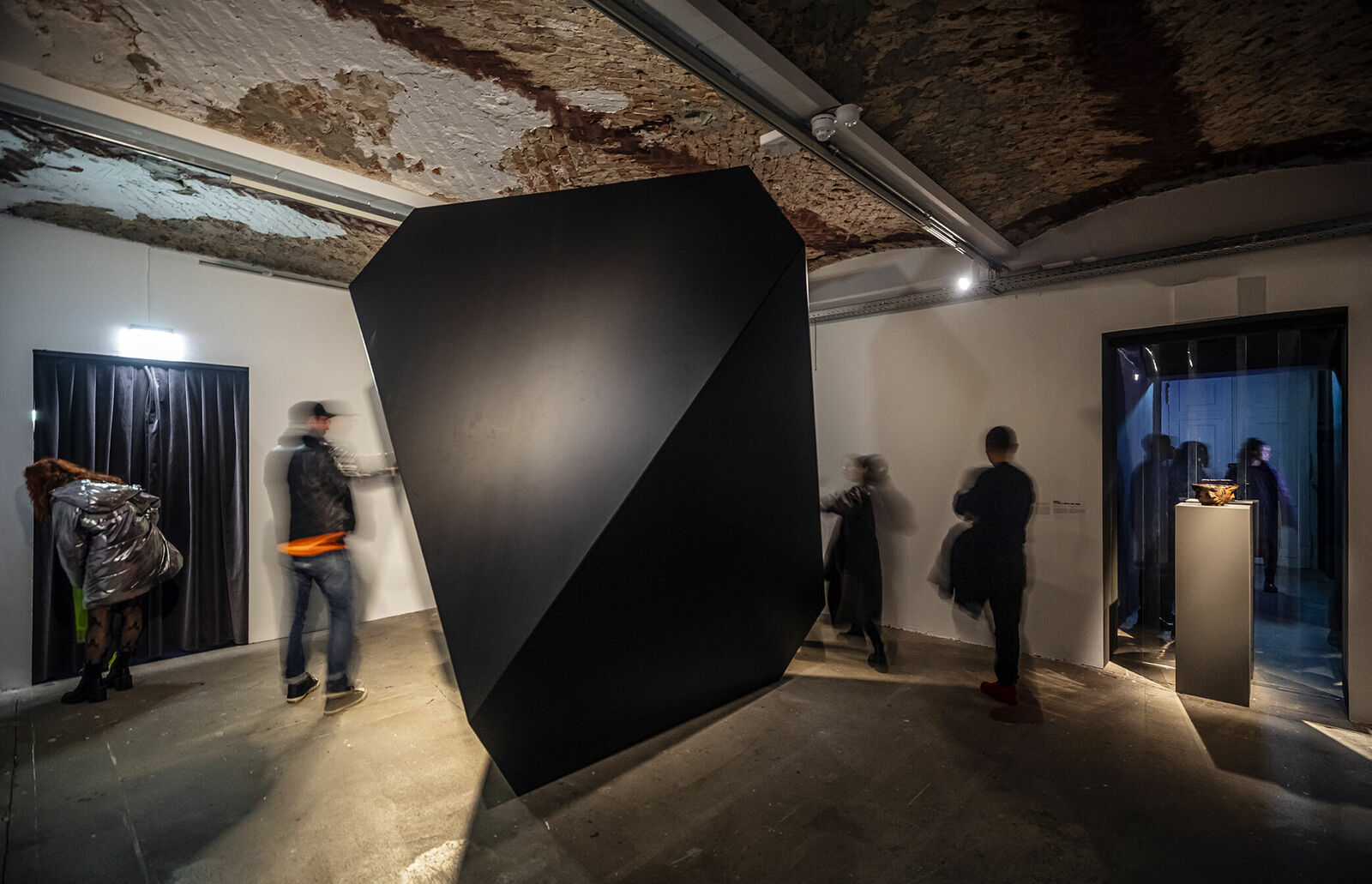 Eine Kunstinstallation in einem Raum mit rustikalem Ambiente. Im Zentrum steht ein großes, monolithisches schwarzes Objekt. Besucher bewegen sich um die Skulptur herum und durch den Raum