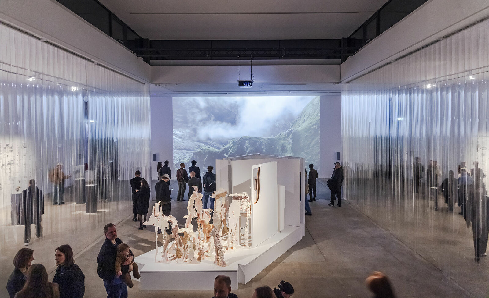 Besucher betrachten Kunstwerke in einem modernen Galerieraum, abgetrennt durch transparente, hängende Trennwände. Im Zentrum steht eine Skulpturenausstellung auf einem weißen Podest, und an der hinteren Wand wird ein großes Landschaftsbild projiziert