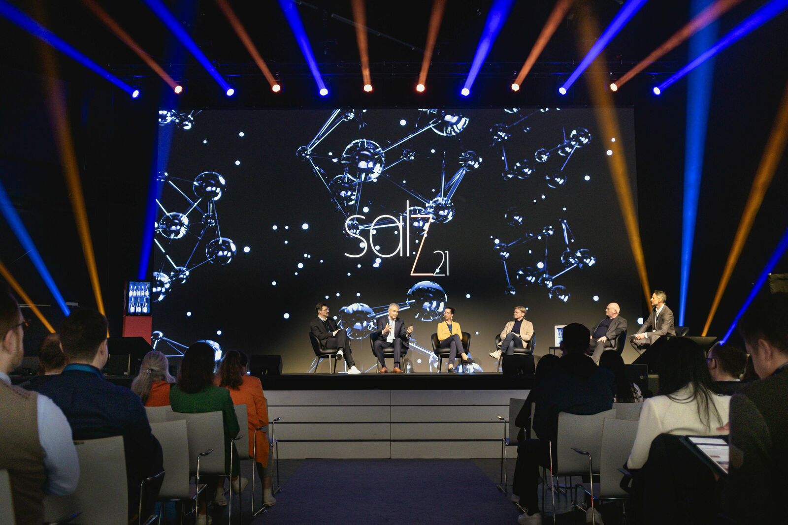Eine Konferenzszene mit sechs Personen auf einer Bühne, vor einem großen Bildschirm mit dem Wort "SALT" und der Zahl "21", umgeben von Sternbildgrafiken.
