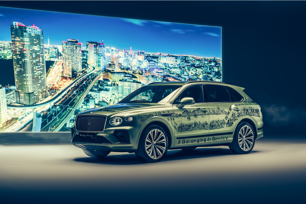 Ein Bentley-SUV vor einem großen Bildschirm, der eine nächtliche Stadtszene zeigt. Das Auto ist auffallend mit einer Skizze der Stadt und dem Hashtag #Belonging4Bentley bedruckt