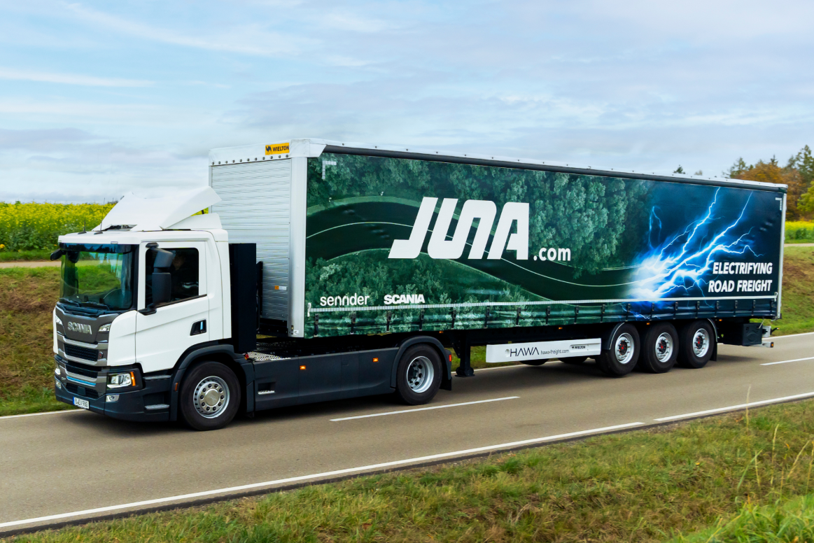 Ein weißer Scania-LKW fährt auf einer Straße neben einem Feld. Der LKW-Anhänger ist mit einer auffälligen Werbung für 'JUNA.com' bedruckt