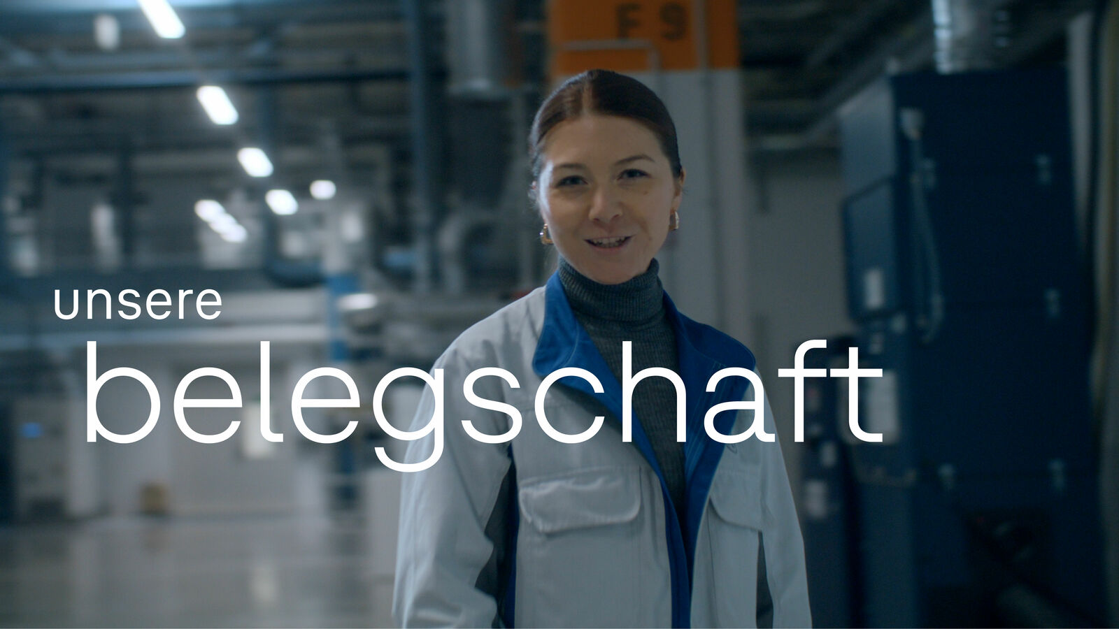 Lächelnde Frau mit zurückgebundenen Haaren, die eine Arbeitskittel und einen blauen Rollkragen trägt, steht in einer industriellen Umgebung. Über ihr erscheint der Text 'unsere Belegschaft'