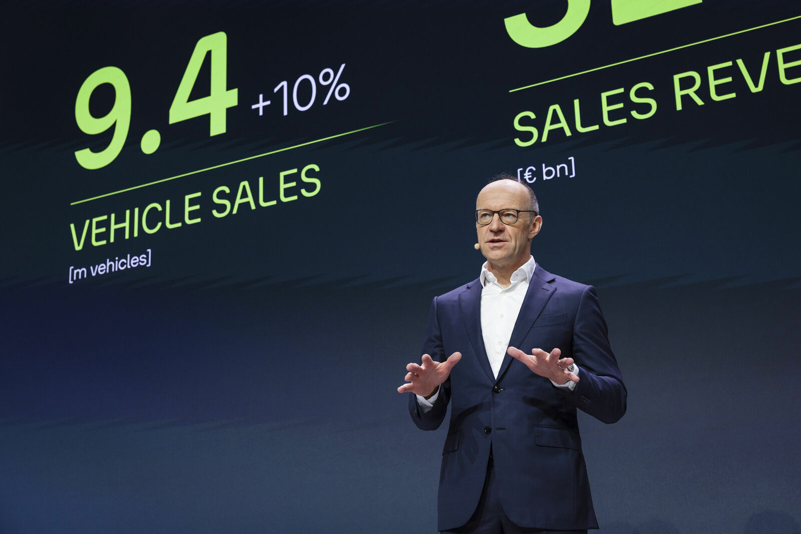 A man on stage presenting vehicle sales and sales revenue figures in front of a large display board.