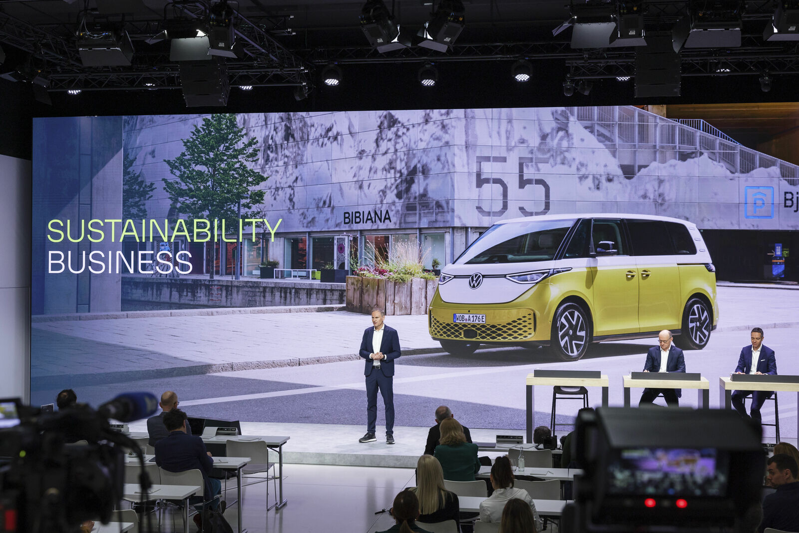 A man presents on stage about sustainability in business featuring an image of a yellow, electrical VW bus.