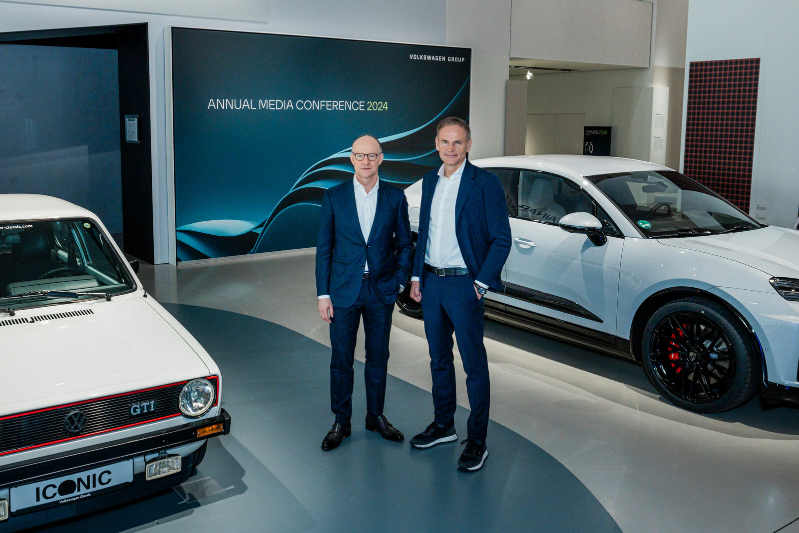 Two men in business attire standing next to a classic and a modern Volkswagen car at a press conference.