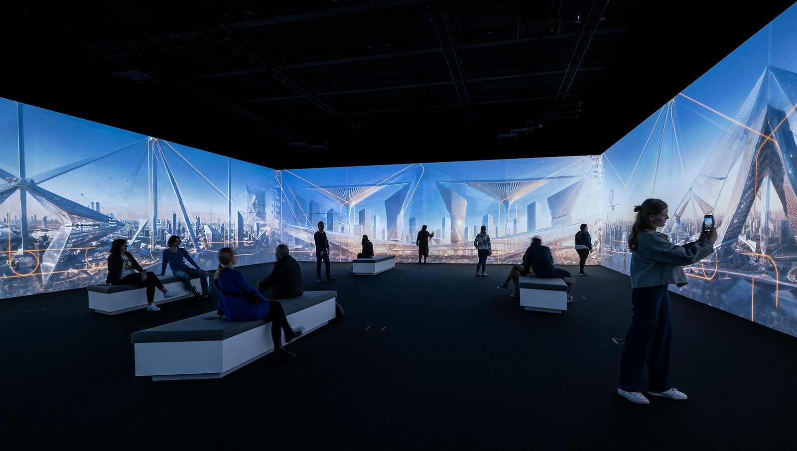 Visitors in a dark exhibit hall surrounded by large-scale illuminated cityscape projections.