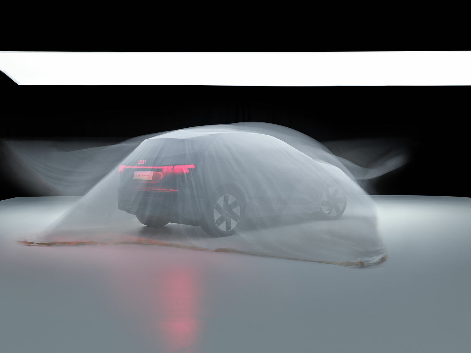 An Audi, covered by a transparent cloth that emphasizes the shape of the car and the red tail lights, stands in a dark room.