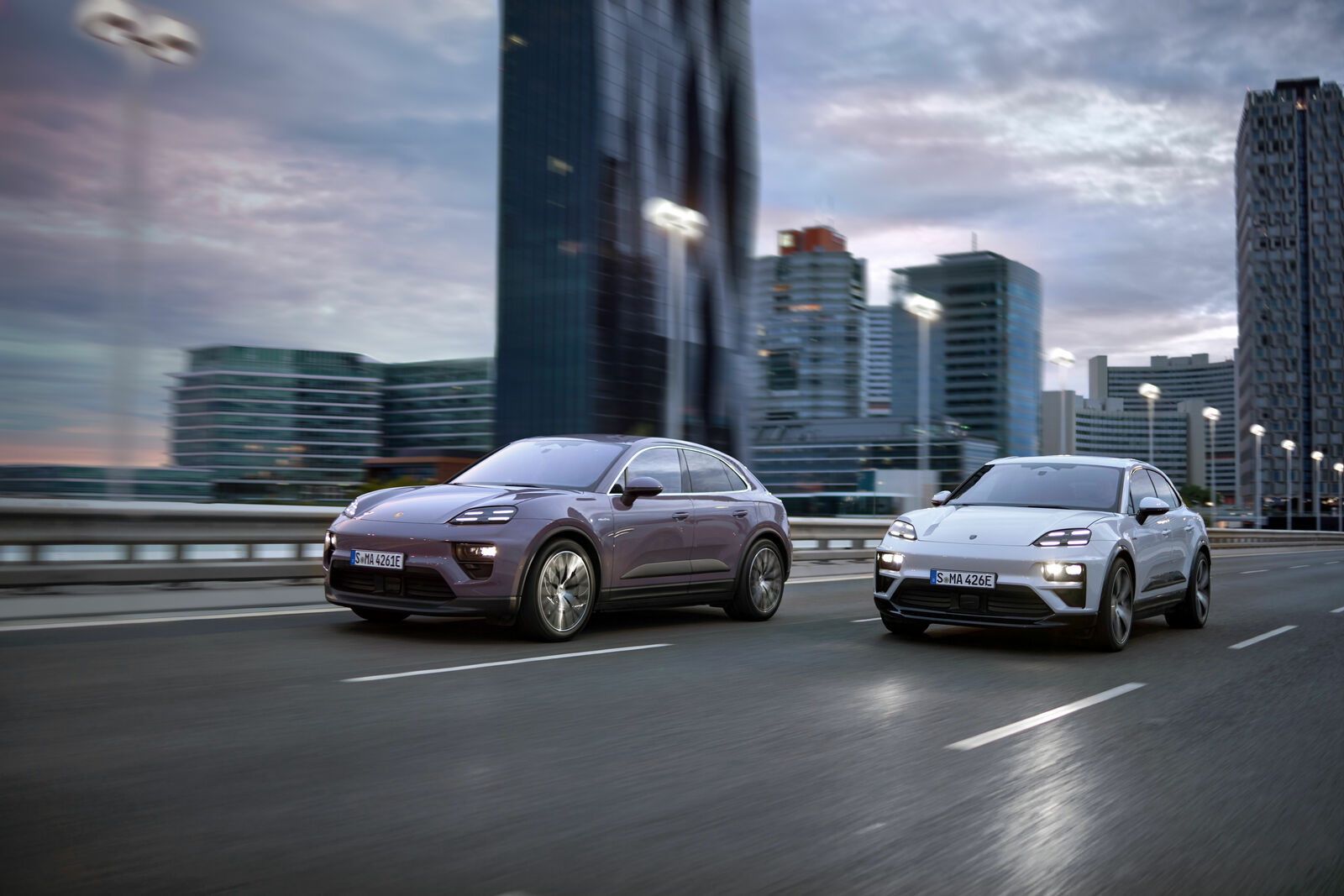 Two modern electric SUVs in lilac and silver drive side by side on a city highway, with skyscrapers and a cloudy sky in the background.