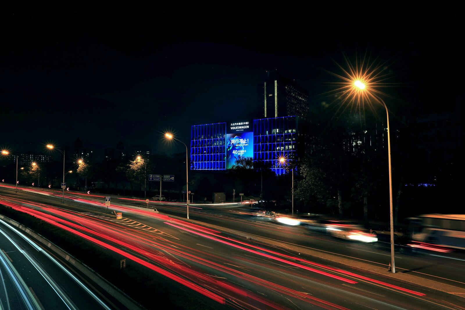 Nachtansicht einer belebten Straße mit Lichtspuren von Fahrzeugen und einem beleuchteten Volkswagen-Gebäude im Hintergrund.