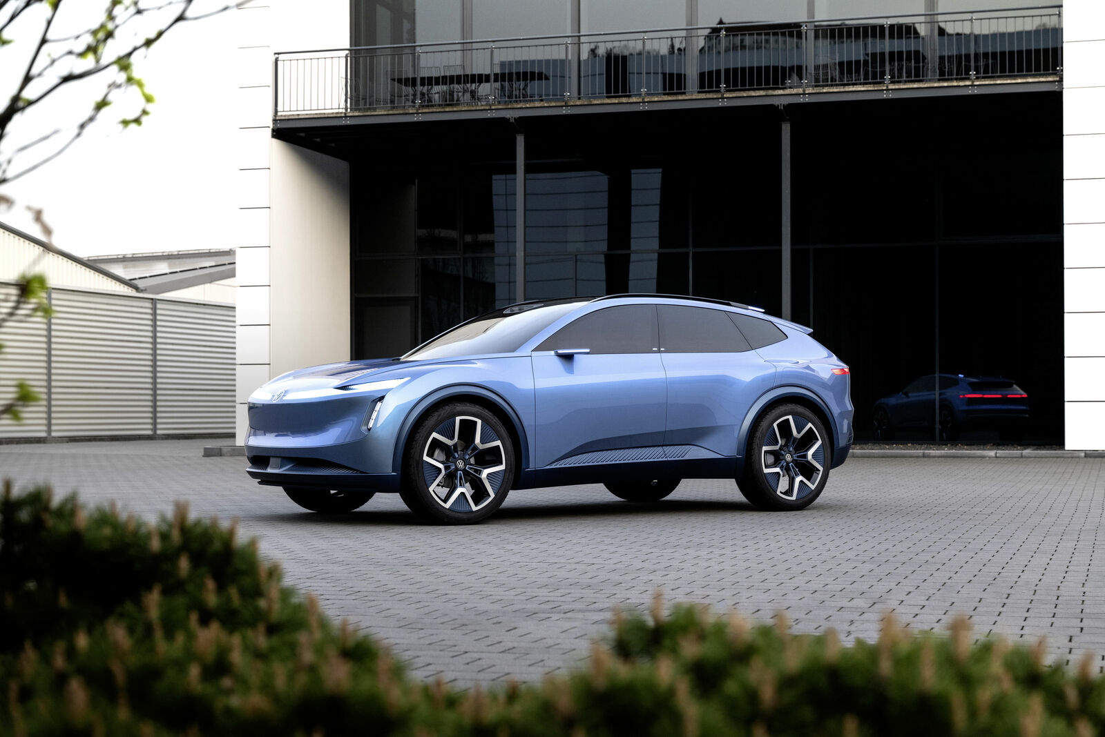An image of a futuristic blue concept car parked on a paved plaza in front of a modern building with large glass facades and metal structures.