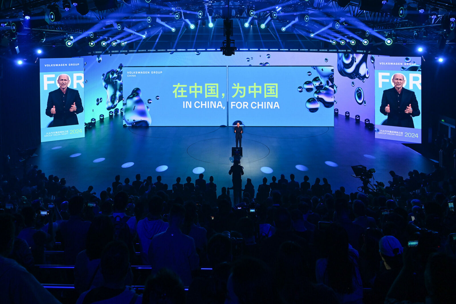 Ein Redner steht in der Mitte einer hell beleuchteten Bühne und spricht vor einem großen Publikum. Der Hintergrund zeigt den Slogan "IN CHINA, FOR CHINA" in Englisch und Chinesisch und hebt den strategischen Fokus der Volkswagen-Gruppe auf den chinesischen Markt hervor.