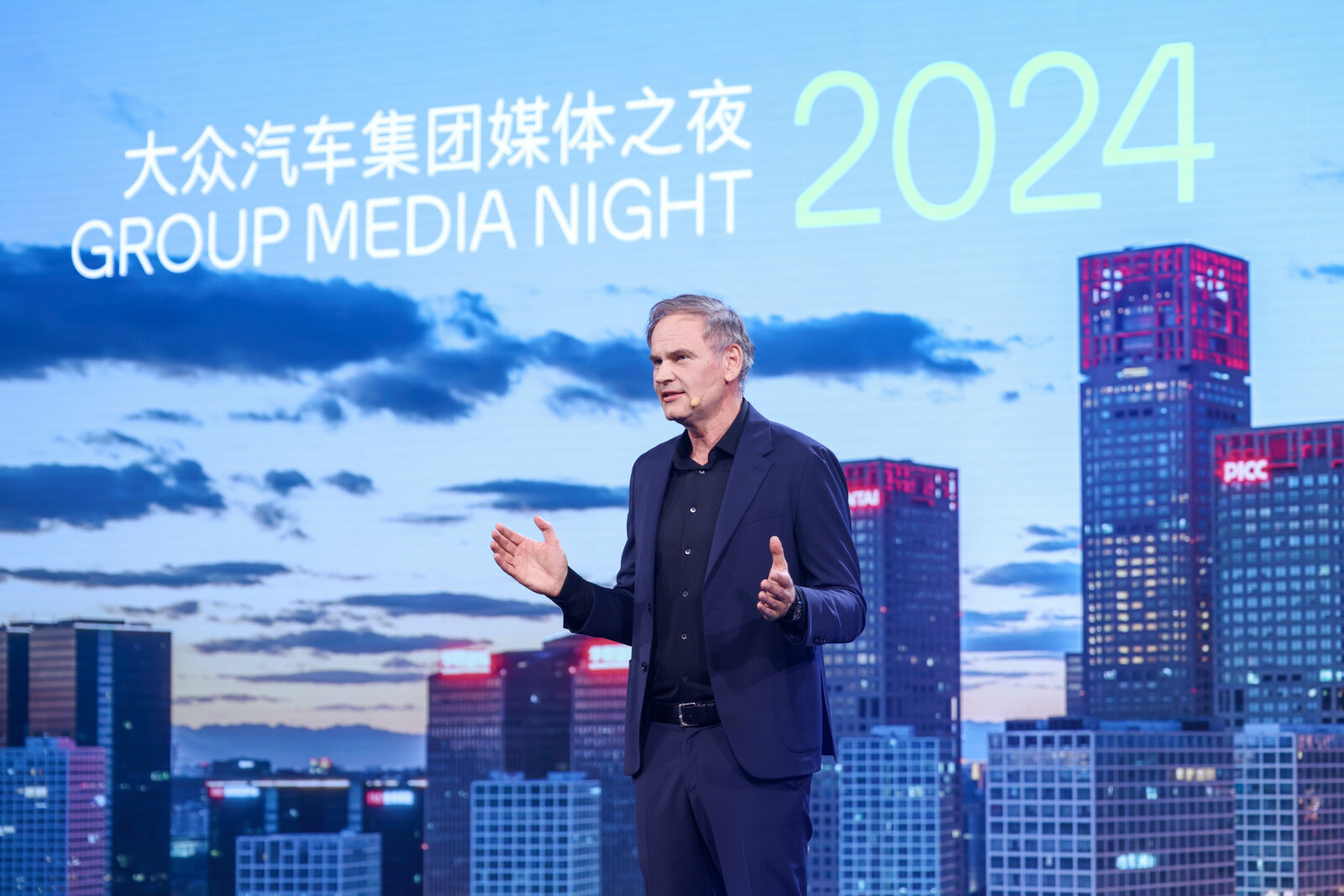 A presenter speaks at the "Group Media Night 2024" event, gesturing with his hands to emphasize his points. Behind him, the screen shows a cityscape with a twilight sky, while the event title and year are prominently displayed in both Chinese and English.