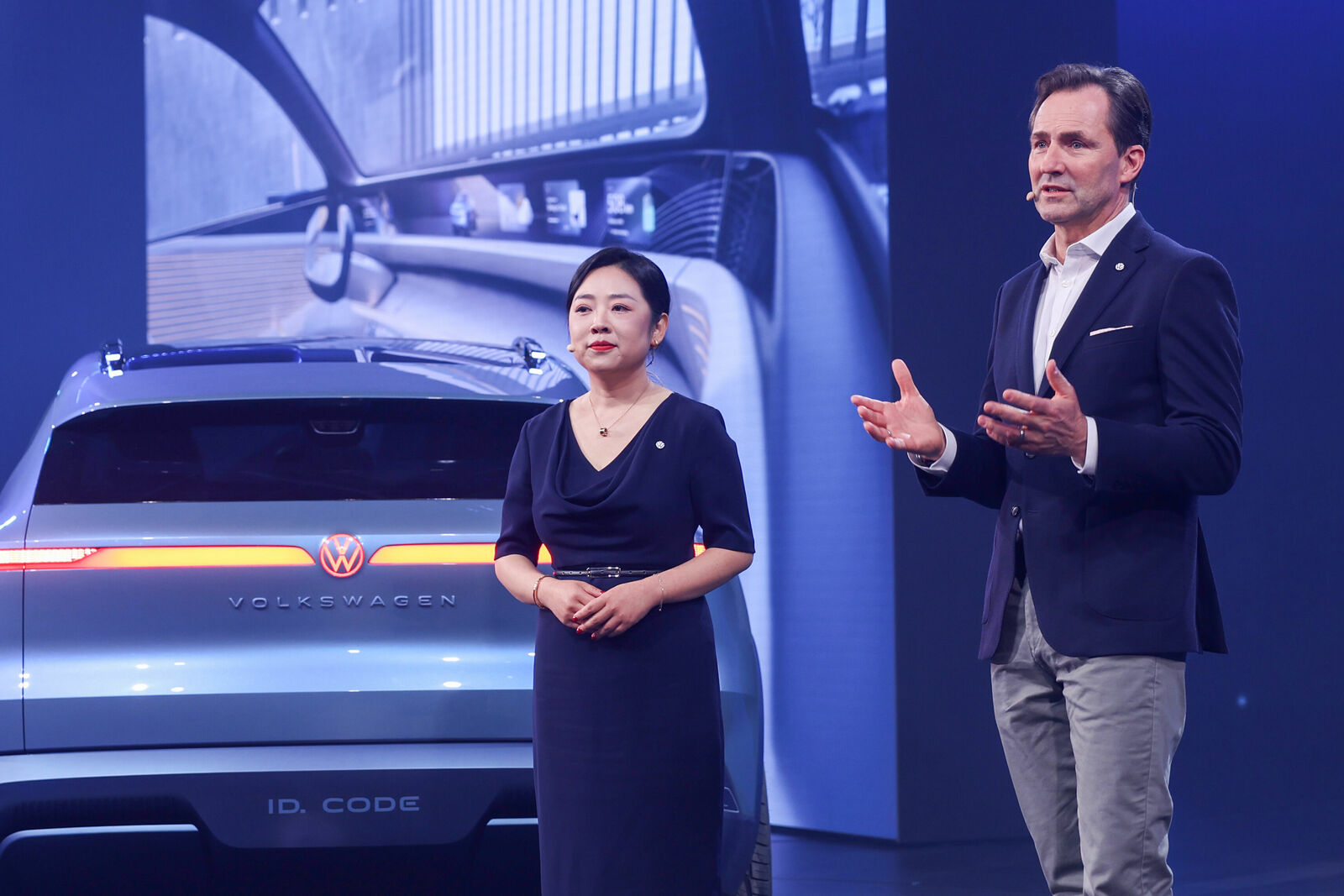 Two presenters, a woman and a man, stand confidently on stage in front of a metallic blue Volkswagen ID. Code concept SUV. They are delivering a presentation about the vehicle, gesturing and speaking to the audience.