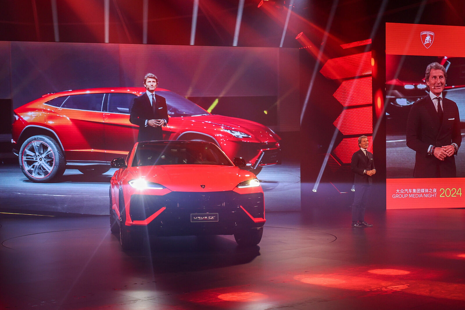 A bright orange Lamborghini Urus S is showcased under vibrant red lighting in a presentation space.