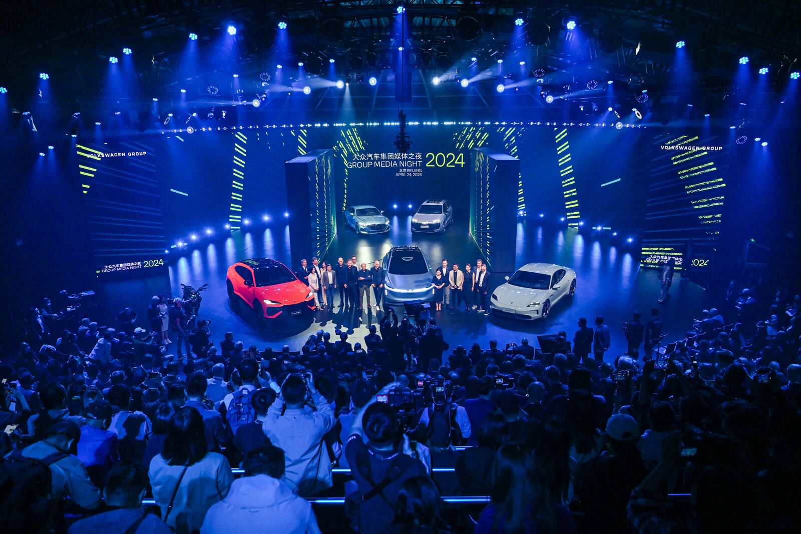 Eine große Menschenmenge besucht die Veranstaltung "Group Media Night 2024", bei der mehrere hochwertige Autos auf der Bühne unter heller blauer Beleuchtung präsentiert werden.